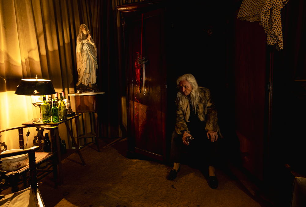 Olwen Fouere's character Rita Concannon drinking while sitting down in her room with a bottle on her hand. The room has a statue of Mary and bottles on a table.